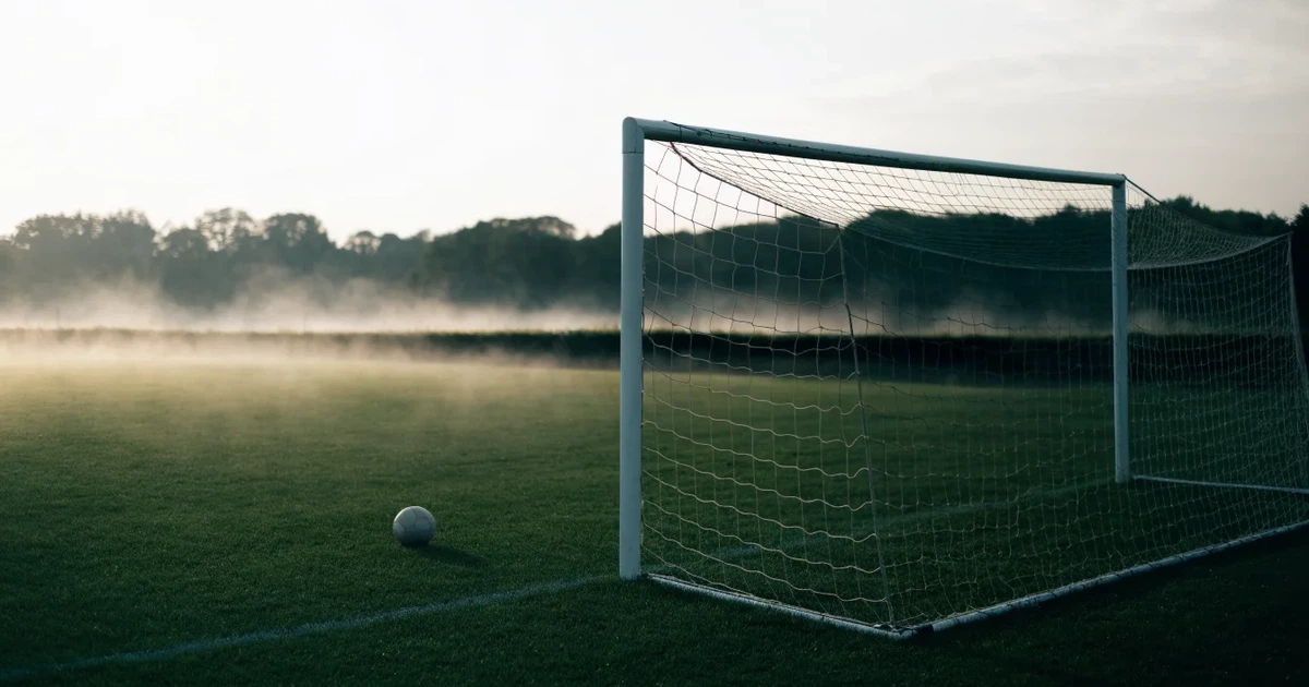 Ein Fußballtor mit gespanntem Netz steht auf einem leeren Rasenplatz, davor ein einzelner Ball, Morgendunst liegt über dem Feld.