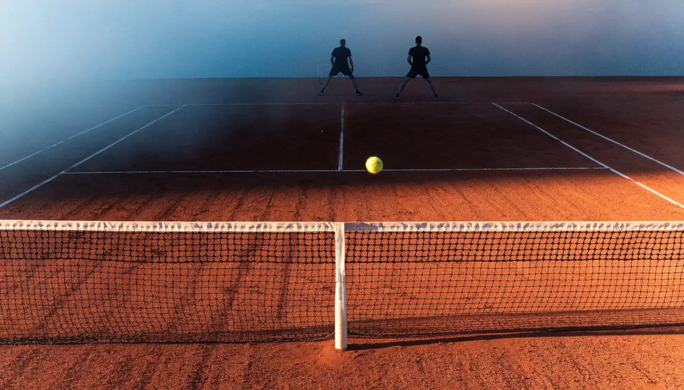 Ein Tennisplatz aus Sand bei Sonnenaufgang, das Netz im Gegenlicht, kein Mensch zu sehen.