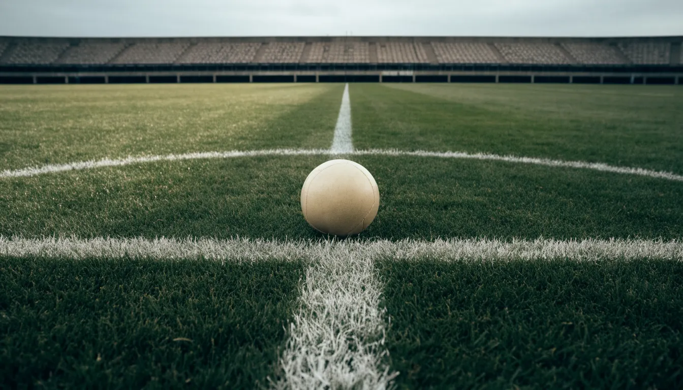 Ein Fußball liegt auf dem Anstoßpunkt eines leeren Rasenplatzes, im Hintergrund eine ruhige Tribüne unter bedecktem Himmel.