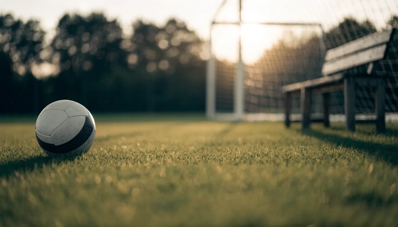 Ein Fußball liegt im Gras am Spielfeldrand, dahinter eine leere Bank und ein Tor im Gegenlicht.