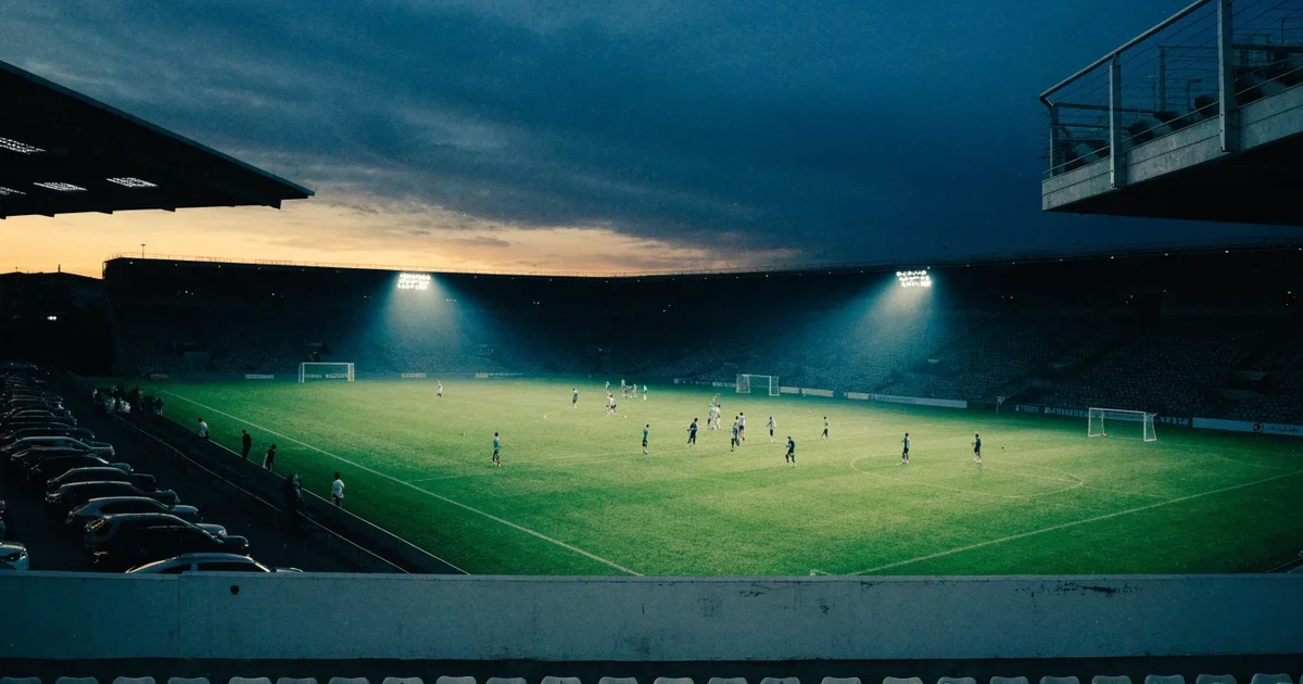 Ein Fußballstadion unter Flutlicht aus großer Entfernung, davor ein dunkler Parkplatz und ein heller Abendhimmel.