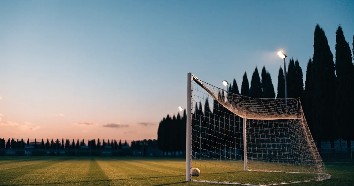 Ein Fußballtor mit Netz auf einem leeren Platz im Abendlicht, der Rasen frisch gemäht, im Hintergrund Zypressen.