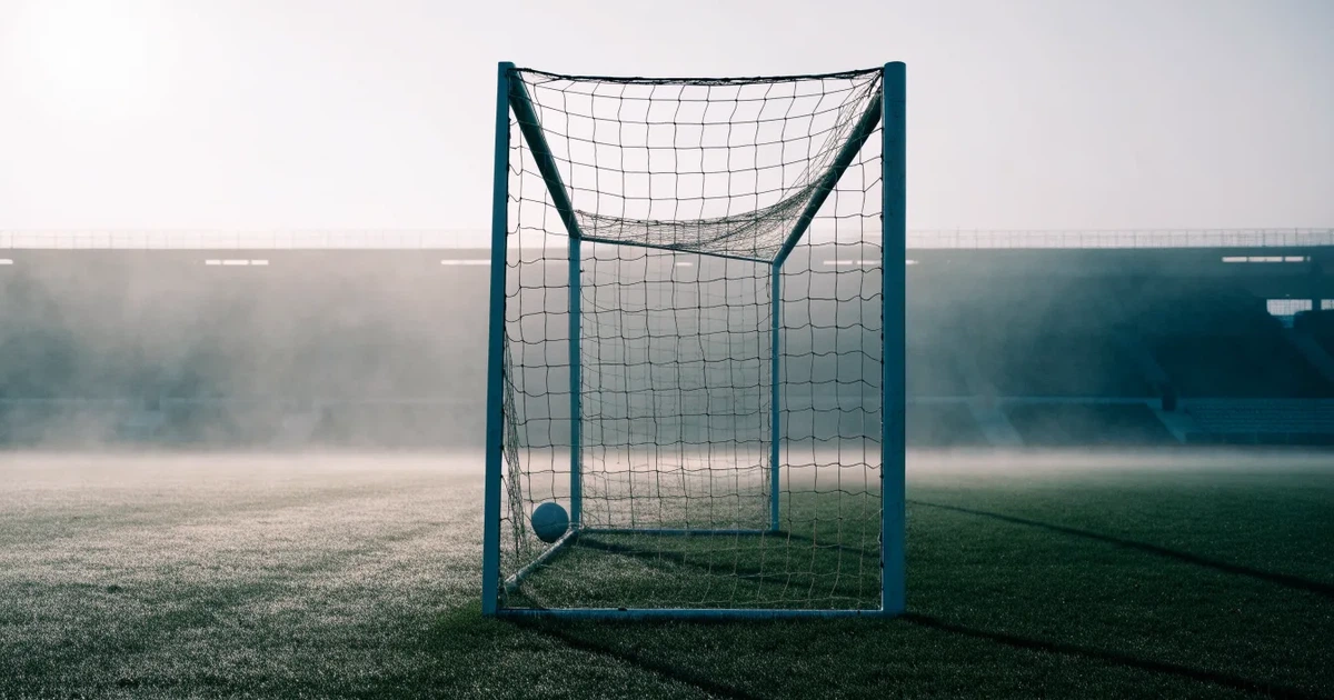 Ein Fußballtor mit Netz steht auf einem leeren Platz im Nebel der Morgendämmerung, der Rasen ist taufeucht.