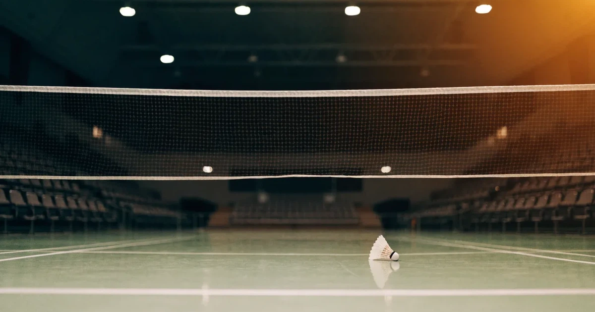 Eine leere Badmintonhalle mit gespanntem Netz und hellen Hallenlichtern, ein Federball liegt auf dem Hallenboden.
