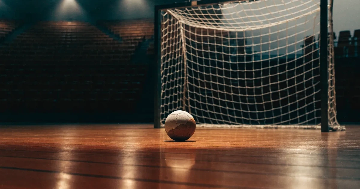 Ein Handball liegt auf dem Hallenboden vor einem Tor mit Netz, die leere Halle wird von Flutlicht beleuchtet.