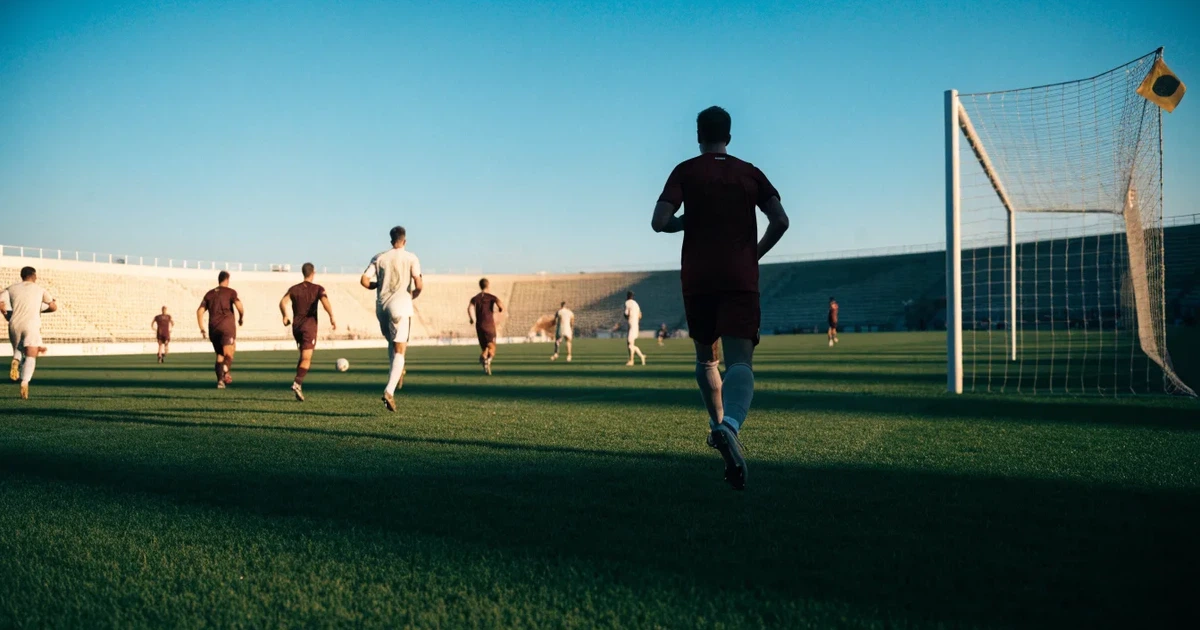 Ein Fußballstadion mit hellen Rängen unter strahlend blauem Himmel, davor ein sonnenbeschienener Rasen mit frisch gemähten Streifen.