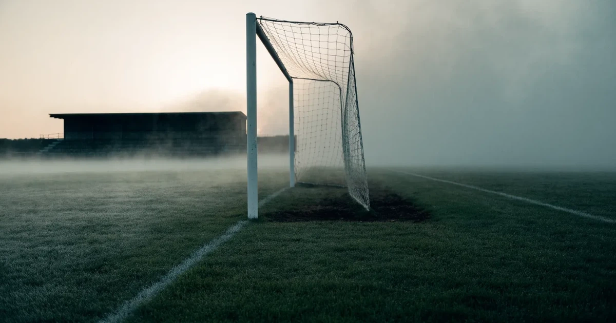 Ein Fußballtor mit Netz steht im Morgennebel auf einem leeren Rasenplatz, im Hintergrund die Silhouette einer kleinen Tribüne.