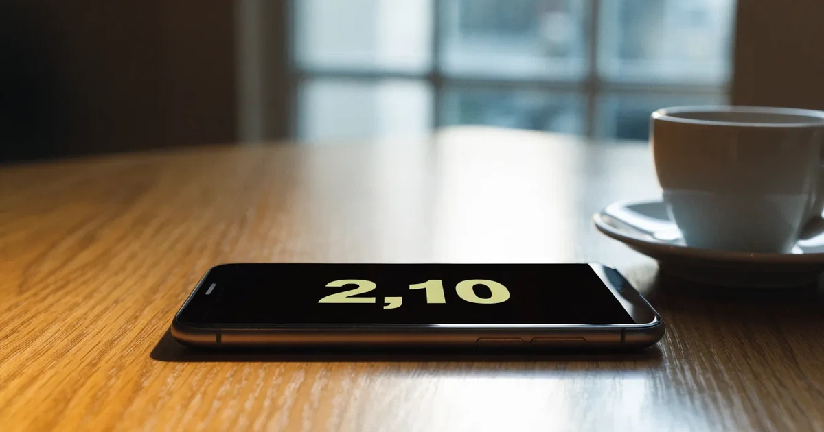 Ein Smartphone mit dunklem Display liegt auf einem Holztisch neben einer Tasse Kaffee, im Hintergrund ein unscharfes Fenster.