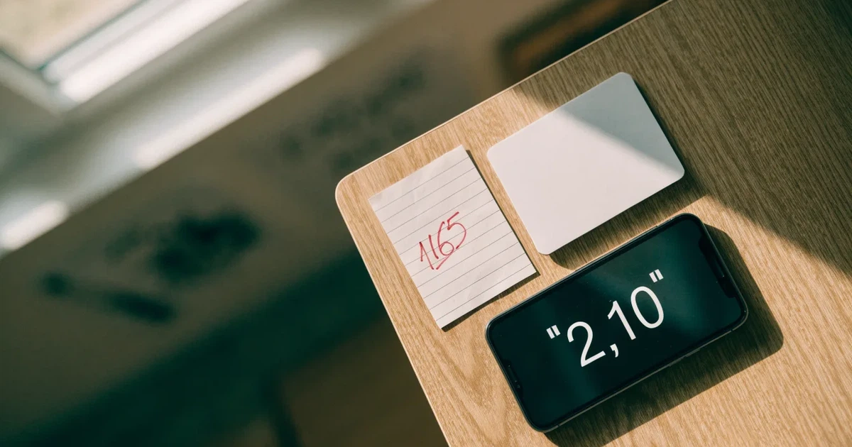 Eine Plastikkarte ohne Aufdruck liegt neben Münzen und einem Smartphone mit dunklem Display auf einem hellen Tisch.