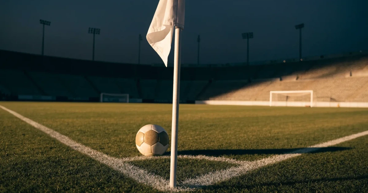 Ein Fußball liegt am Viertelkreis an der Eckfahne eines Rasenplatzes, im Hintergrund leere Tribünen in warmem Abendlicht.