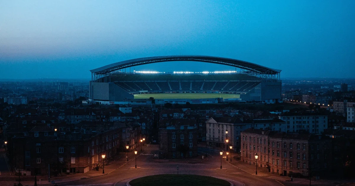 Ein Fußballstadion in der Dämmerung aus der Ferne, die Flutlichter gerade eingeschaltet, davor ein Kreisverkehr mit Laternen.